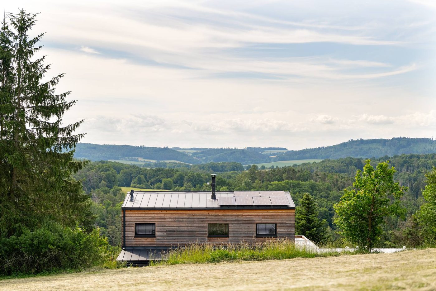 The house from the meadow behind it, low against the treeline, with the valley and the hills beyond